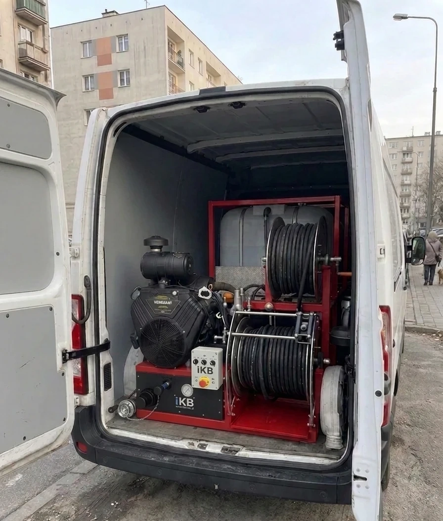 Biały bus z agregatem wuko w środku – zbiornik wody i bębny z wężami wysokociśnieniowymi – zaparkowany przy osiedlu bloków